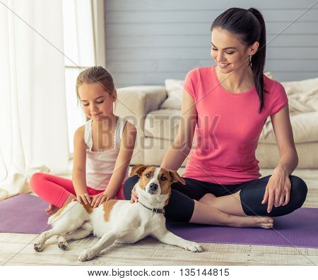 Beautiful young mother and her cute little daughter are patting their dog and smiling while sitting in lotus pose on yoga mat at home