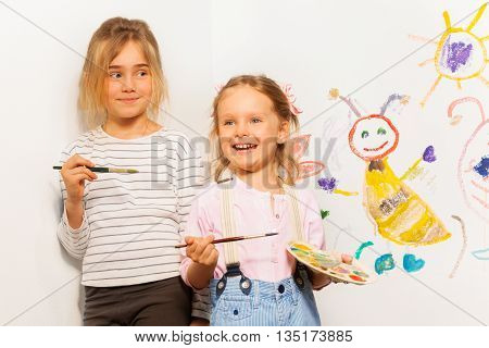 Two smiling painters, girls with pallet and brushes drawing funny picture on the wall
