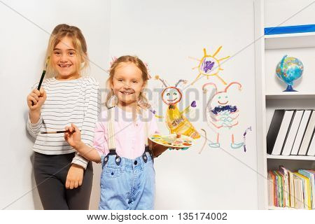 Two happy girls, young painters with brushes and pallet, drawing funny picture at the wall