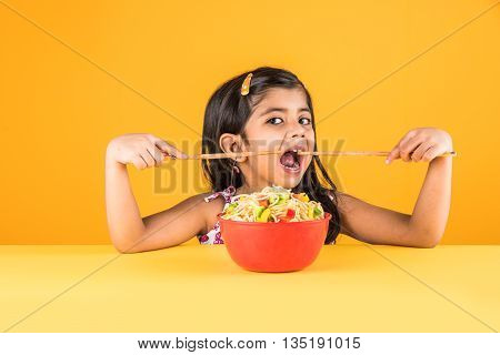 Happy Asian child eating delicious noodle, small indian girl eating noodles with chopsticks in red bowl, over colourful background