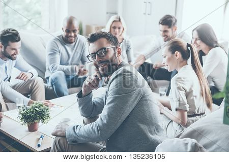 Feeling confident in his team. Group of cheerful young business people sitting at the office desk together and discussing something while young man looking over shoulder and smiling