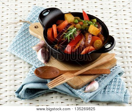 Delicious Homemade Colorful Vegetables Ragout with Striped Eggplant Carrots Potatoes Red Bell Pepper and Green Pea in Black Iron Stewpot with Wooden Fork and Spoon closeup Blue Napkin on Wicker background