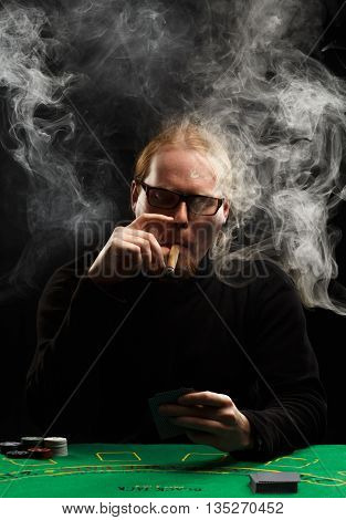 Young red-haired bearded man in glasses and black pullover with a cigar and cards sitting at poker table covered with green cloth in a puff of smoke.