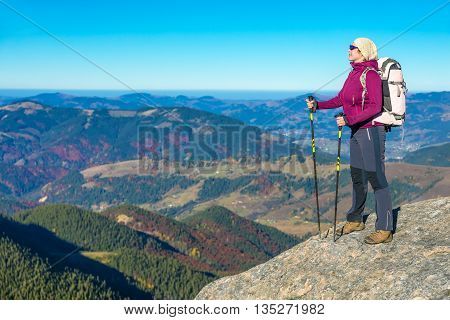 Person with Hiking Backpack Sporty Clothing and Trekking Poles Staying on High Rock with Mountain View with Autumnal Colors Forest and Blue Sky on Background