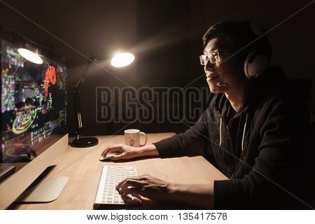 Happy handsome asian male gamer playing computer game at the table in dark room