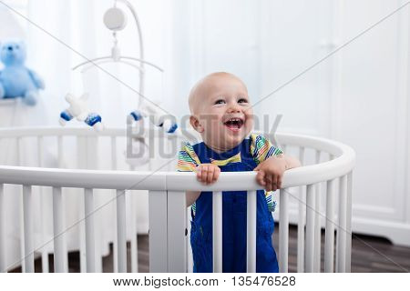 Cute laughing baby standing in a white round bed. White nursery for young children. Little boy learning to stand in his crib. Toys for infant cot. Smiling child playing with toy bear in sunny bedroom.