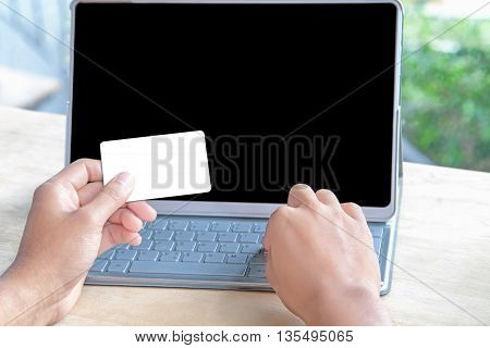 Close-up of person holding empty credit card or business card and forefinger touching keyboard of a blank screen computer tablet. on a wooden table under morning sunlight. online shopping concept.
