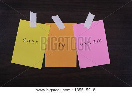 Word quotes of dare to dream on sticky color papers against wooden textured background.