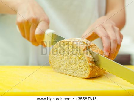 Cutting slices from loaf of bread sitting on yellow surface, hand holding large knife.