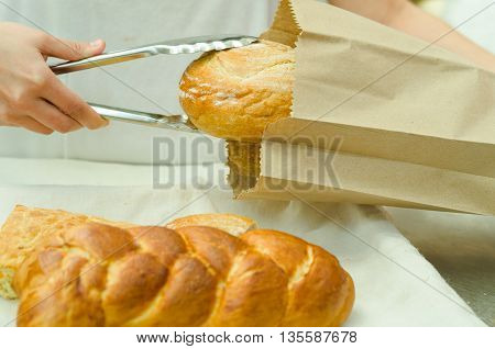 Bakery worker placing loaf of bread inside brown paper bag using large silver tweezers.