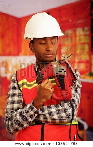 Young engineer carpenter wearing square pattern flanel shirt with red safety vest, holding jigsaw smiling to camera.