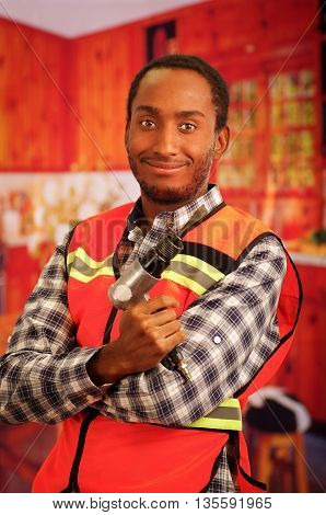 Young engineer carpenter wearing square pattern flanel shirt with red safety vest, holding handheld electric power tool smiling to camera.