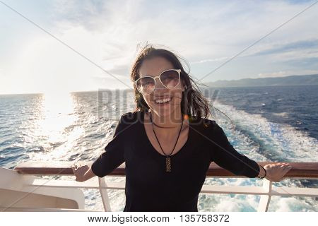 Young fashionable woman in black dress standing on shipboard enjoying sun on her face.Summer vacation,open sea, cruise. Luxury lifestyle,successful woman on earned cruise.Europe summer holidays