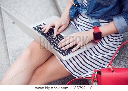 Always connected. Pleasant young woman sitting on the footsteps and typing while using laptop