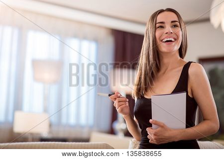 Look at me. Gorgeous young woman with wide smile holding a book and a pen while standing and looking aside