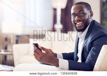 Nice time. Pleasant mature afro American man looking at camera while holding a smart phone and sitting on the couch