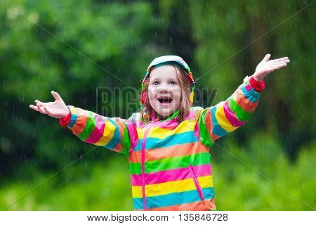 Little girl playing in rainy summer park. Child with rainbow umbrella waterproof coat and boots jumping in puddle in the rain. Kid walking in autumn shower. Outdoor fun by any weather