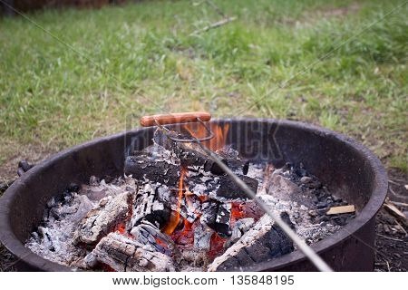 Hot dog on a metal cooking stick cooking over a outdoor camp fire.