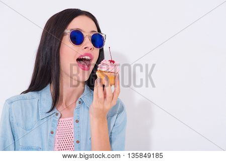 Sweet day. Positive joyful beautiful woman holding cupcake and going to eat it while standing isolated on white background