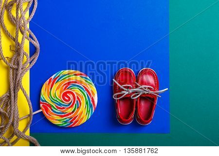 Small red boat shoes near big multi-colored lollipop and rope on colored background. Top view. Copy space frame