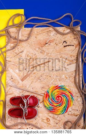 Small red boat shoes near big multi-colored lollipop and rope on wooden desk with colored background. Top view Frame. copy spase.