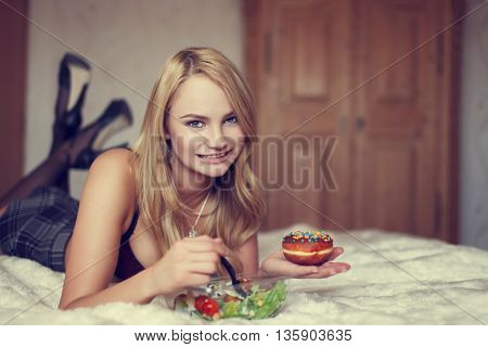 Sexy blonde woman eating salad and dounut on bed teeth smile