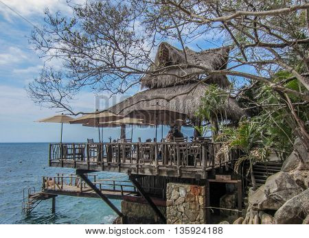 Thatched-roof cliff-side restaurant with umbrellas, tables and chairs hanging over blue ocean with sky of white puffy clouds.