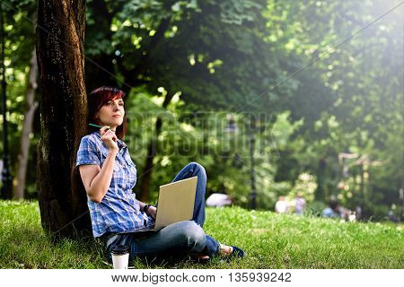 Beautiful young freelancer woman using laptop sitting under the tree in the park.Happy smiling girl working online.Studying and learning using notebook computer.Freelance workbusiness people concept.