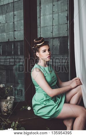 Portrait of the beautiful fasion young woman in cyan green dress with beauty make up, creative hairstyle on brown wooden background. Style photo image of the girl princess, studio shot. Sensual, romantic female sitting at the windowsill.