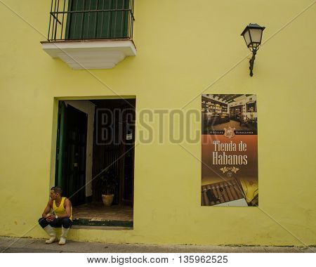 HAVANA - CUBA JUNE 19, 2016: A man sits on the stoop of his home which has been recently painted bright yellow. A poster selling cigars is posted on the wall.