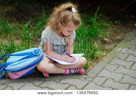 The little schoolgirl sits having crossed legs on a grass and something writes to a notebook. Girlie has a thoughtful look. Near the girl her satchel lies. Back to school.