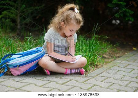 Schoolgirl of elementary school sits having crossed legs on a grass and something writes to a notebook. Girlie has a thoughtful look. Near the girl her satchel lies. Back to school.