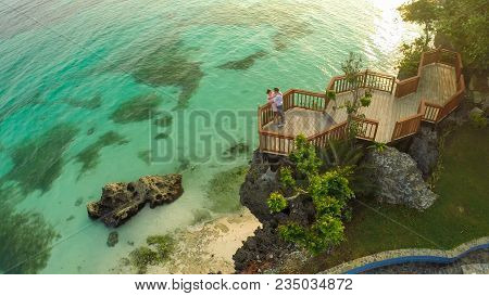 A Beautiful Light Green Coast With Reefs And A Loving Couple On The Balcony Above The Beach. The Bea