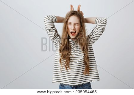 Indoor Portrait Of Good-looking Young European Woman Winking, Holding Fingers Behind Head As If Mimi