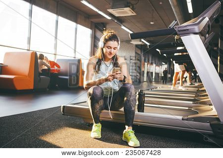 Beautiful Young Caucasian Girl Sportswoman Sitting, Resting After Training On Treadmill Against The 
