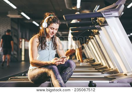 Beautiful Young Caucasian Girl Sportswoman Sitting, Resting After Training On Treadmill Against The 