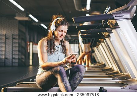 Beautiful Young Caucasian Girl Sportswoman Sitting, Resting After Training On Treadmill Against The 