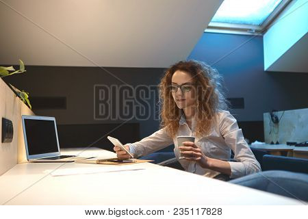 Attractive Young Businesswoman With Wavy Hair Reading Text Message On Cell Phone, Having Coffee Brea
