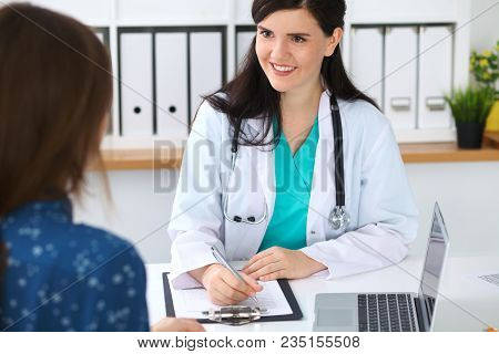 Beautiful Female Doctor And Patient Having A Talk While Sitting At The Desk. Physician Writes Prescr