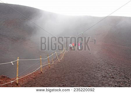 People Walk Along The Crater Of Mount Etna. Black Volcanic Earth And Thick Fog On Mount Etna. Place 