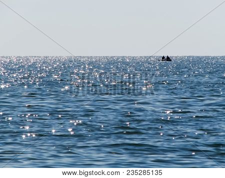 A Blue Sea With Small Waves And Sun Glares, A Lonely Boat With Two Men In The Distance, And A Clear 