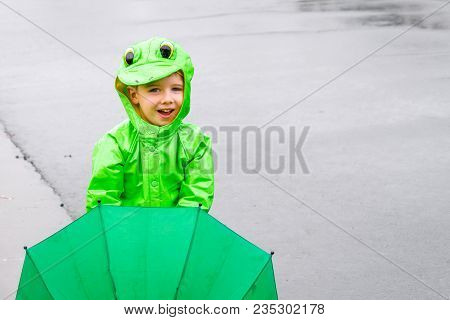 A Little Boy Stands On A Street In A Frog Raincoat And Holding A Green Umbrella In Front Of Him On A