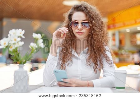 Horizontal Shot Of Beautiful Curly Woman With Confident Look, Wears Sunglasses And White Blouse, Hol