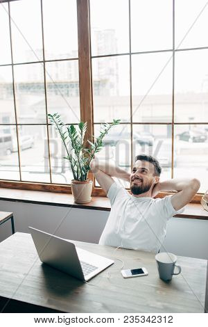 Vertical Picture Of Bearded Man Sitting At The Table And Relaxing. He Is Near Tbig Window. Guy Is Re