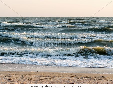 Dark Sea Waves With White Foam Near A Sandy Beach At Sunset