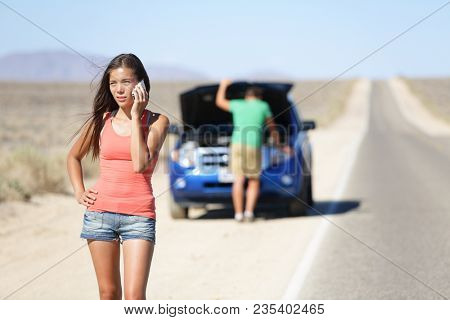 Car breakdown - woman phone calling auto service help. Neutral or negative expression on woman on mobile smart phone, while man trying to fix the car. Young couple on road highway in California, USA.