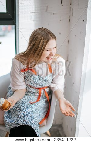 Blonde Happy Young Woman In Mottled Apron Having Cupcake And Laughing Sitting Near The Window