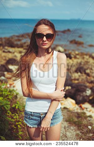 Portrait Of Pretty Young Woman In Sunglasses Standing On Coastline With Ocean On Background
