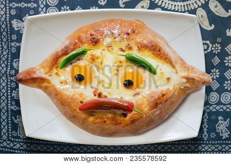 Georgian Dish Adjarian Khachapuri With Eggs, With Sad Face On  Plate, Blue Tableclothe Background,