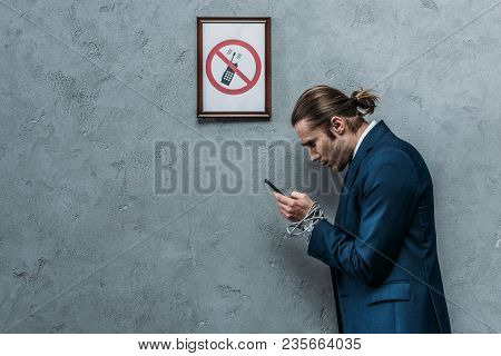 Depressed Businessman In Suit With Hands Tied With Wire Using Smartphone Under Restricted Smartphone
